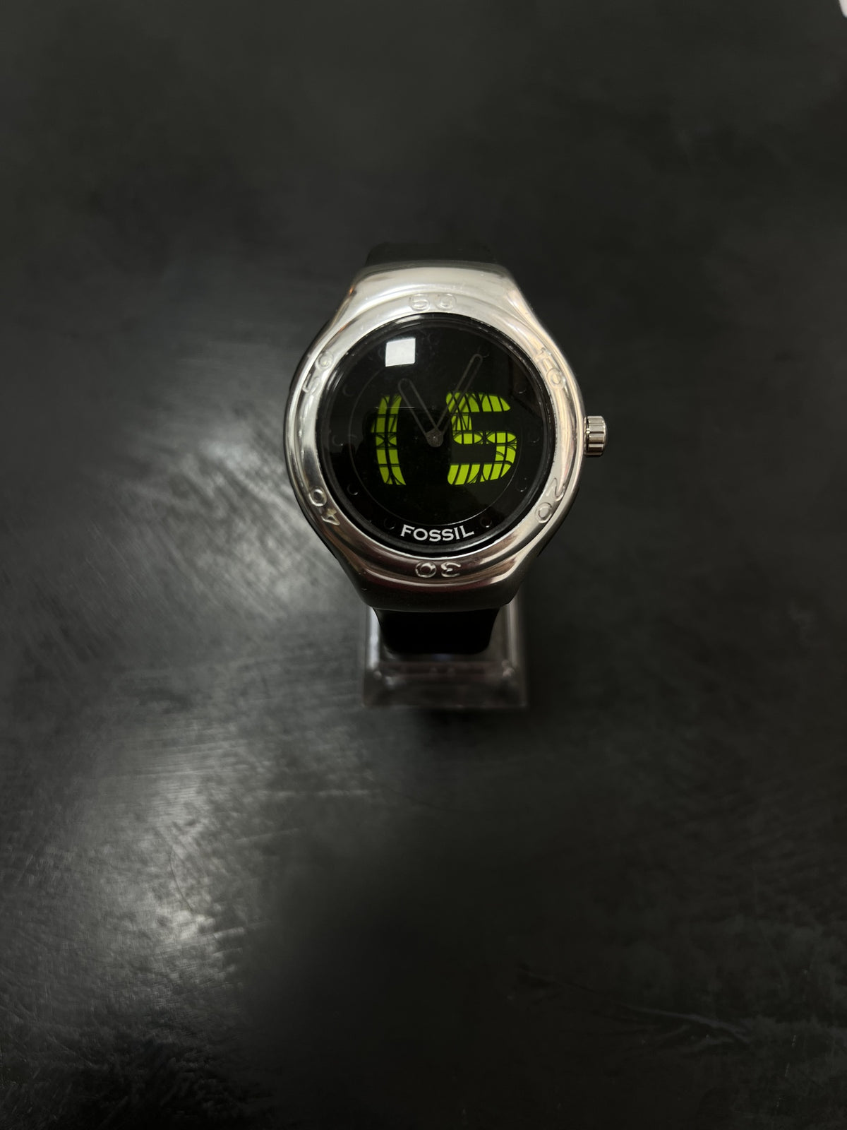 Reloj Fossil Big Tic con caja plateada, correa negra y dígitos verdes sobre fondo oscuro.